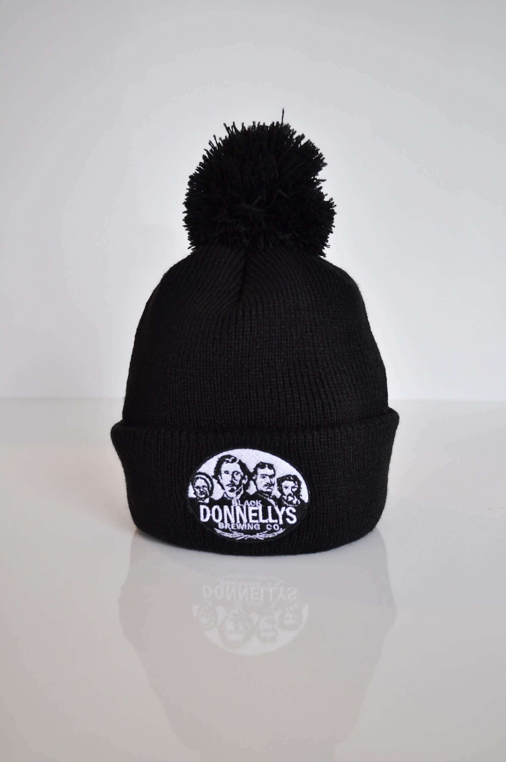 A Black Donnellys Brewing Company Toque Style Winter Hat Topped With Pom-pom And Black And White Company Logo On The Folded Brim.